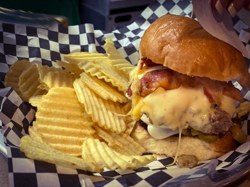 A juicy burger with melted cheese, bacon, and pickles, served with a side of ridged potato chips in a checkered basket.