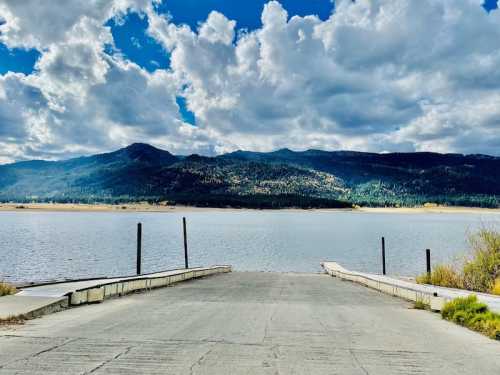 A serene lake view with a concrete ramp leading to the water, surrounded by mountains and fluffy clouds.