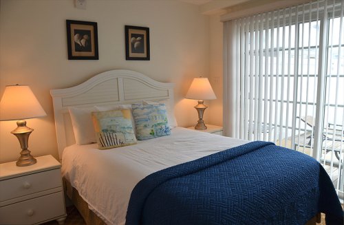 Cozy bedroom with a white bed, blue blanket, bedside lamps, and artwork on the walls, featuring natural light from windows.