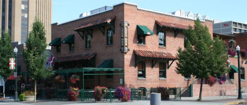 A brick restaurant with green awnings, surrounded by trees and colorful flowers, in an urban setting.