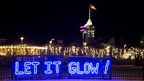 A brightly lit sign reading "LET IT GLOW!" in blue neon, with festive lights and a tower in the background at night.