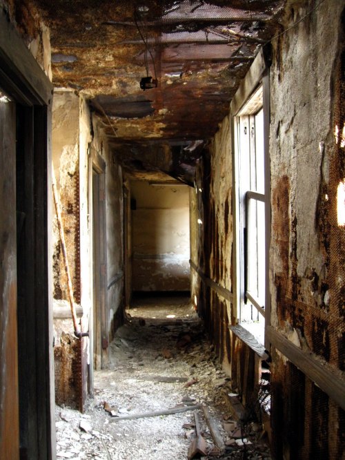A dilapidated hallway with peeling walls, debris on the floor, and dim light at the far end.