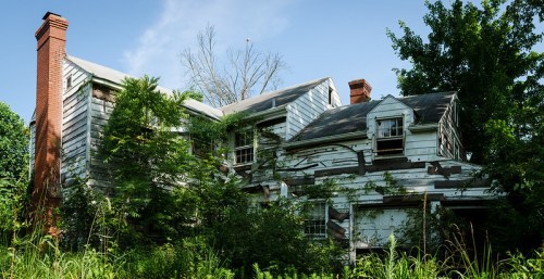Abandoned house overgrown with vegetation, featuring peeling paint and a weathered exterior under a clear blue sky.