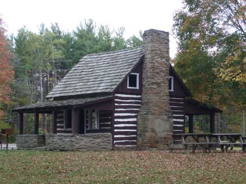 A rustic log cabin with a stone chimney, surrounded by trees and fallen leaves in a serene outdoor setting.