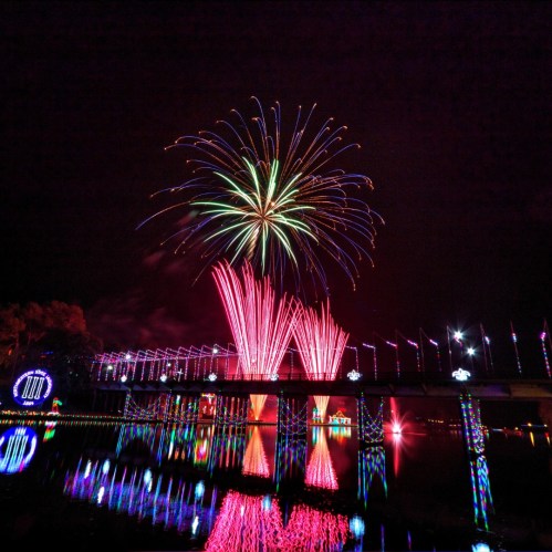 Colorful fireworks burst over a dark sky, reflecting in a calm body of water, with illuminated decorations along the shore.