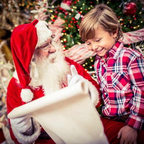 Santa Claus smiles at a young boy in a festive setting, as they share a moment by a decorated Christmas tree.