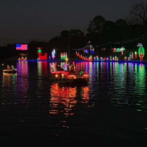 Boats decorated with colorful lights on a dark lake, reflecting festive decorations and an American flag in the background.