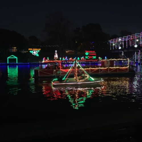 A decorated boat with colorful lights sails on a dark river, reflecting festive decorations from the shore.