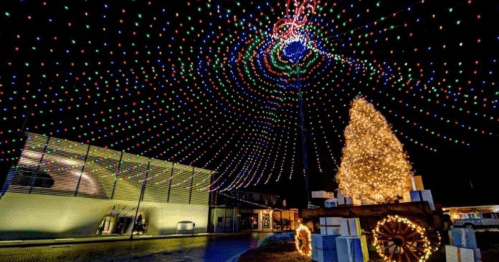Colorful holiday lights create a spiral pattern above a decorated Christmas tree and presents in a festive outdoor setting.
