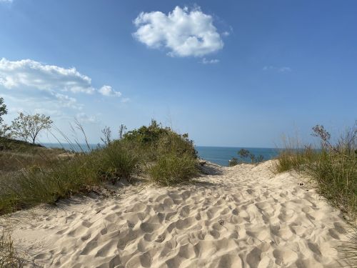 A sandy path leads to a serene beach with gentle waves under a blue sky and scattered clouds.