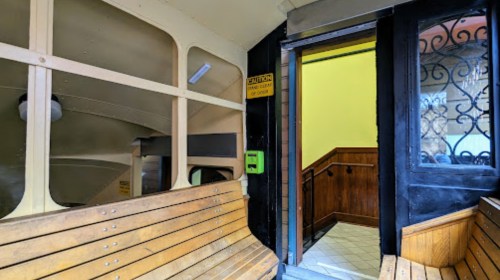 Interior of a bus with wooden benches, leading to an open door and staircase, with a yellow wall visible.