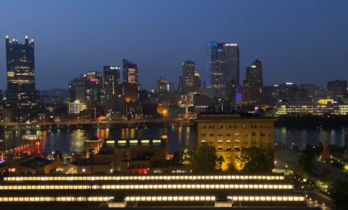 City skyline at night with illuminated buildings and a river reflecting lights in the foreground.