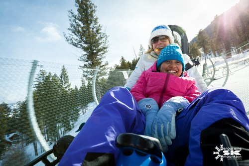 A woman and a child smile while riding a snowy mountain lift, surrounded by trees and bright sunlight.