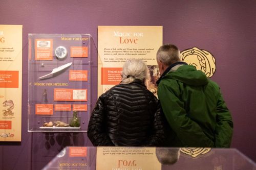 Two visitors examine an exhibit titled "Magic for Love" in a museum, surrounded by informative panels.
