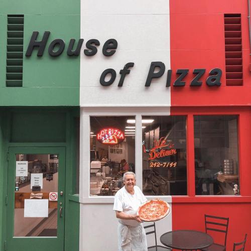 A chef stands outside the "House of Pizza," holding a large pizza in front of a colorful building.