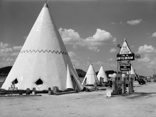 Black and white image of teepees and a vintage gas station sign, with clouds in the background.