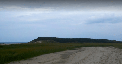 A sandy path leads to a grassy area with a distant hill and cloudy sky over a calm beach.