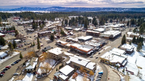 Aerial view of a snowy town with shops, trees, and mountains in the background under a cloudy sky.