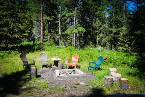 A cozy outdoor fire pit area with colorful chairs, surrounded by trees and a hammock in a lush green forest.