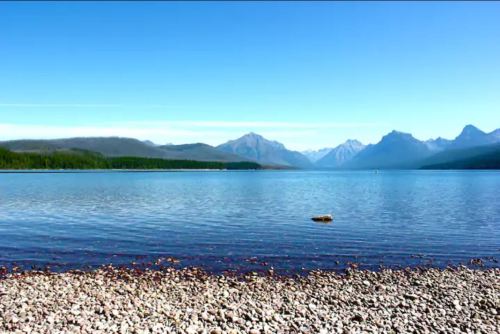 A serene lake surrounded by mountains, with a clear blue sky and a rocky shoreline in the foreground.