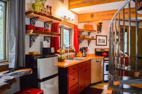 Cozy kitchen with wooden shelves, red accents, a spiral staircase, and modern appliances in a rustic setting.