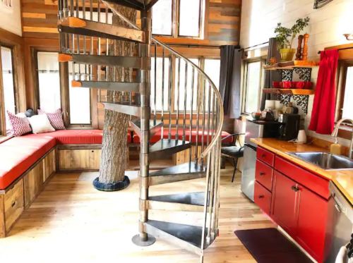 A cozy interior featuring a spiral staircase, wooden accents, a tree, and a red kitchen with a seating area.