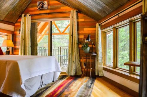 Cozy bedroom with wooden walls, large windows, and a colorful rug, featuring a balcony view of nature.