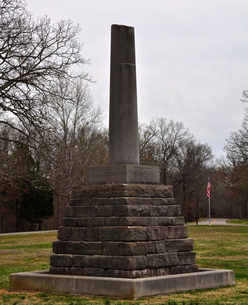 A tall stone monument on a stepped base, surrounded by trees and grass, with a flag in the background.