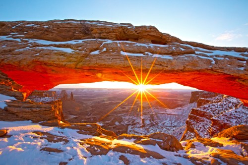 A stunning sunrise shines through a natural rock arch, illuminating snowy landscapes and distant rock formations.