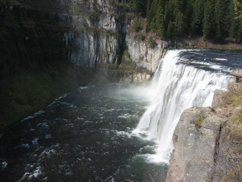 A waterfall cascading over a rocky cliff into a river, surrounded by lush green trees and mist.