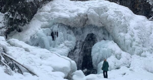 A person stands in front of a large frozen waterfall surrounded by snow-covered rocks and trees.