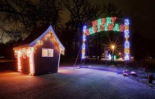 A festive scene featuring a small cabin and a colorful "Merry Christmas" arch illuminated with lights at night.