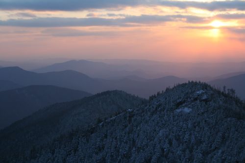 A scenic view of snow-covered mountains at sunset, with layers of hills fading into the distance under a cloudy sky.