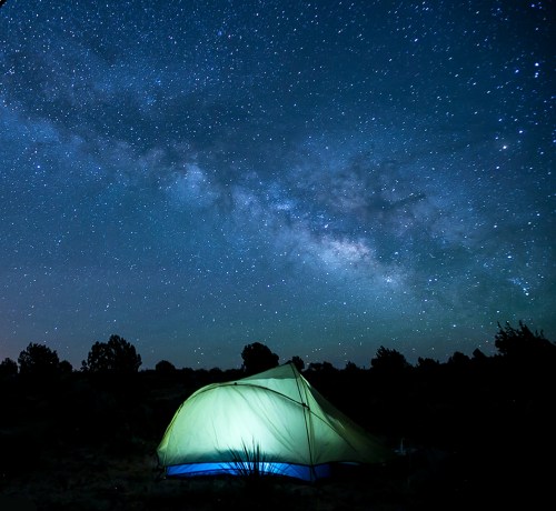 A glowing tent under a starry night sky filled with the Milky Way.