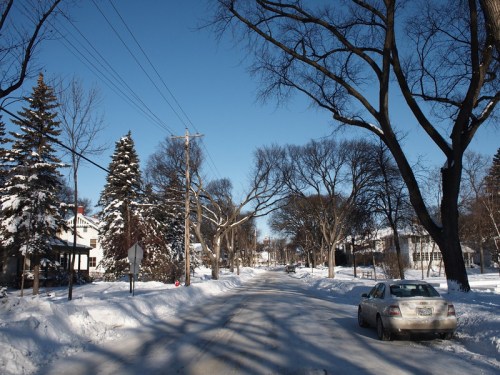 A snowy street lined with trees and houses, with a parked car on the side under a clear blue sky.