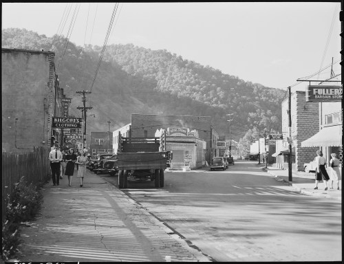 Black and white photo of a small town street with shops, parked cars, and people walking, set against a mountainous backdrop.