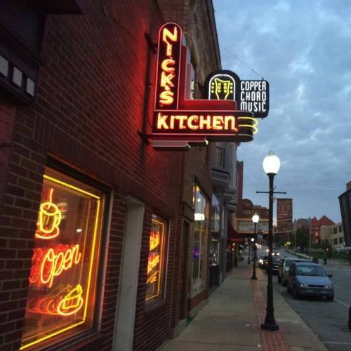 Neon sign for Nick's Kitchen illuminated at night, with nearby streetlights and storefronts visible.