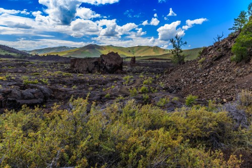 A rocky landscape with sparse vegetation, mountains in the background, and a bright blue sky with scattered clouds.