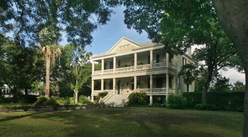 A large, two-story yellow house with a porch, surrounded by trees and a grassy lawn.