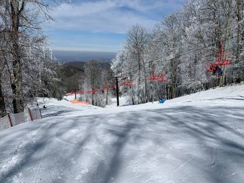 A snowy ski slope with chairlifts in the background, surrounded by trees and a clear blue sky.