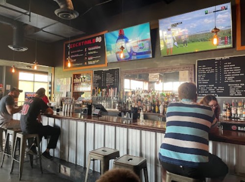 A lively bar scene with patrons sitting at the counter, two TVs showing sports, and a variety of drinks displayed.