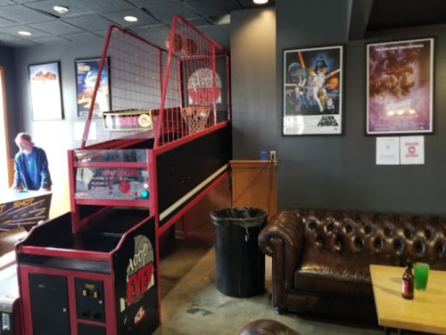 A basketball arcade game in a casual lounge area with a leather couch and posters on the wall.