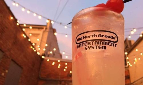 A refreshing cocktail with a cherry garnish, set against a backdrop of string lights and brick walls.