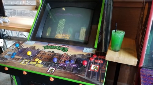 An arcade machine featuring "Teenage Mutant Ninja Turtles" with a green drink beside it on a wooden table.