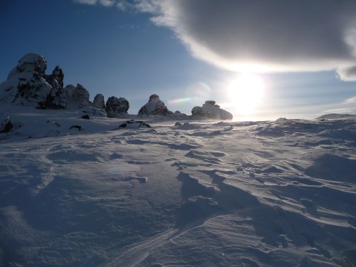 A snowy landscape with rocky formations under a bright sun and a partly cloudy sky.