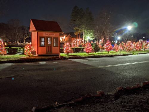 A festive scene at night featuring a small orange building and decorated trees with colorful lights along a road.