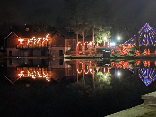 A festive scene at night with illuminated reindeer, colorful lights, and reflections on a calm lake.