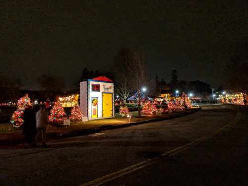 Colorful holiday decorations line a dark street, featuring lit Christmas trees and a whimsical, brightly painted structure.