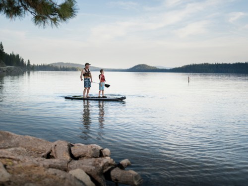 A man and a child stand on a paddleboard in a calm lake, surrounded by trees and mountains in the background.