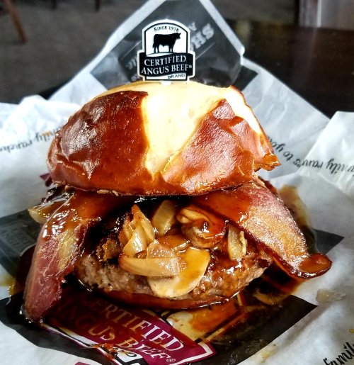 A juicy burger with bacon and sautéed onions on a pretzel bun, topped with a Certified Angus Beef logo.